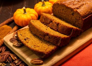 Sliced pumpkin bread loaf sitting on wooden cutting board with pecan nuts and cinnamon spices